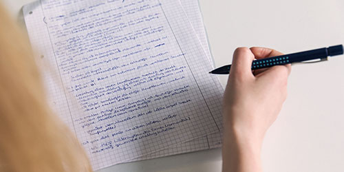 eine Hand hält einen Stift, daneben ein handgeschriebener Text auf einem karierten Blatt